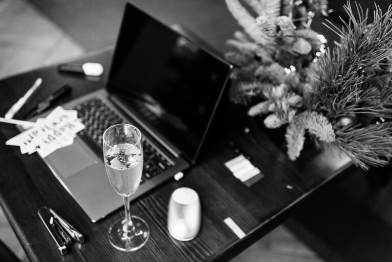 A retro styled black and white photo of a laptop with a glass of champaign nearby and gift tag, with a christmas tree in the background